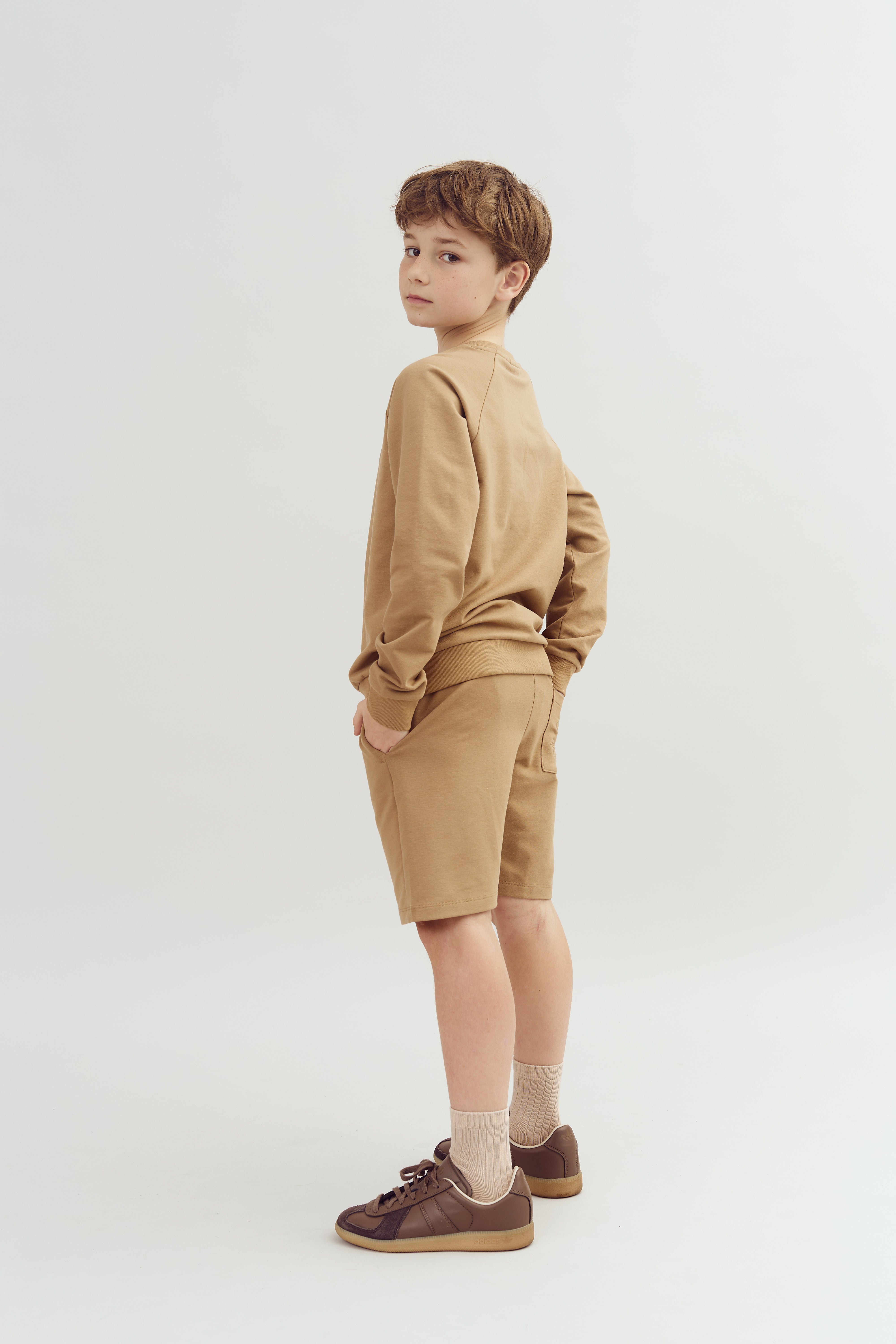 A young boy stands with hands in his pockets, wearing a tan sweatshirt and shorts, brown sneakers, and beige socks. The background is a plain, neutral white.