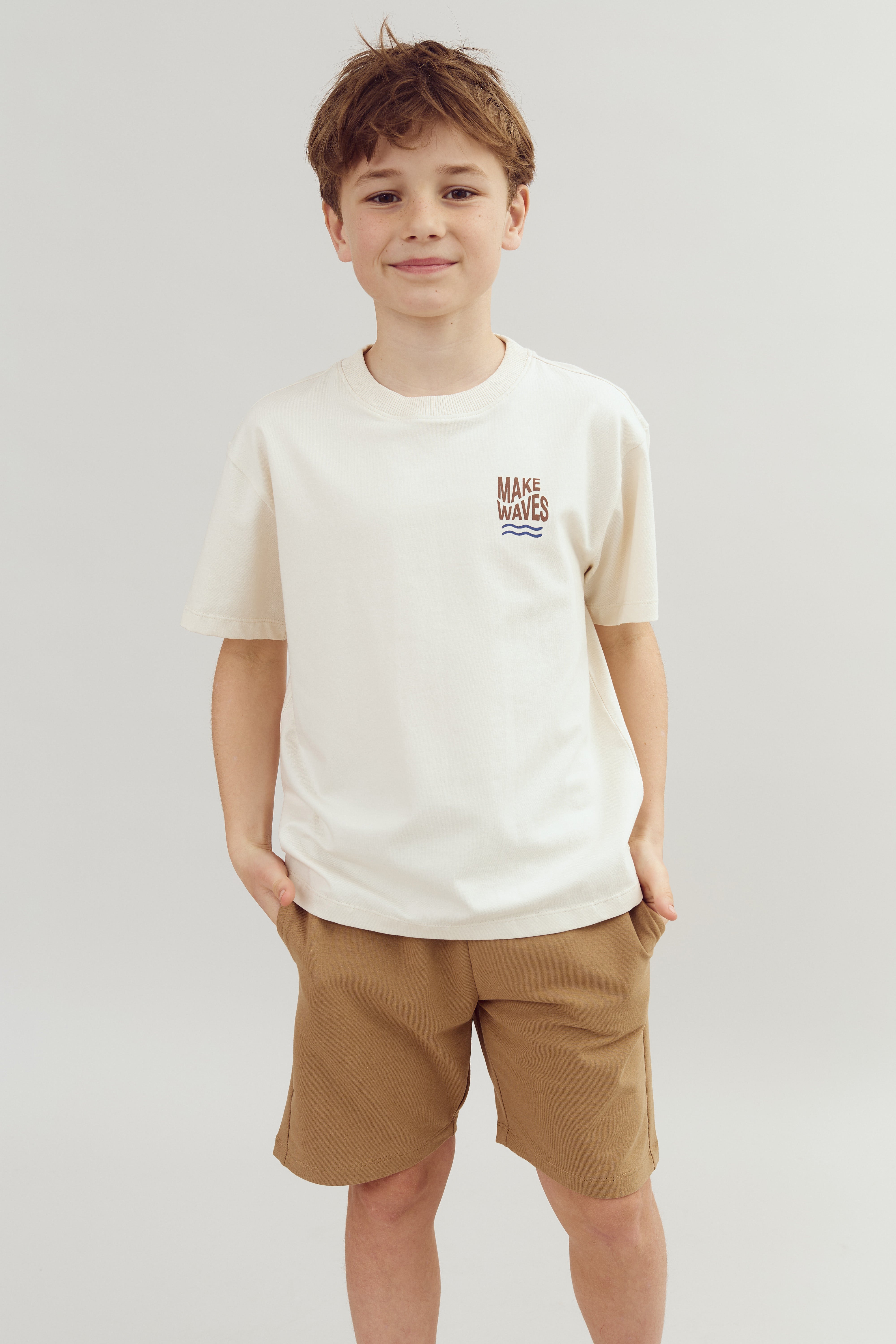 A child stands confidently, wearing a white shirt with "MAKE WAVES" text, and brown shorts. They have hands in pockets against a neutral, plain backdrop.