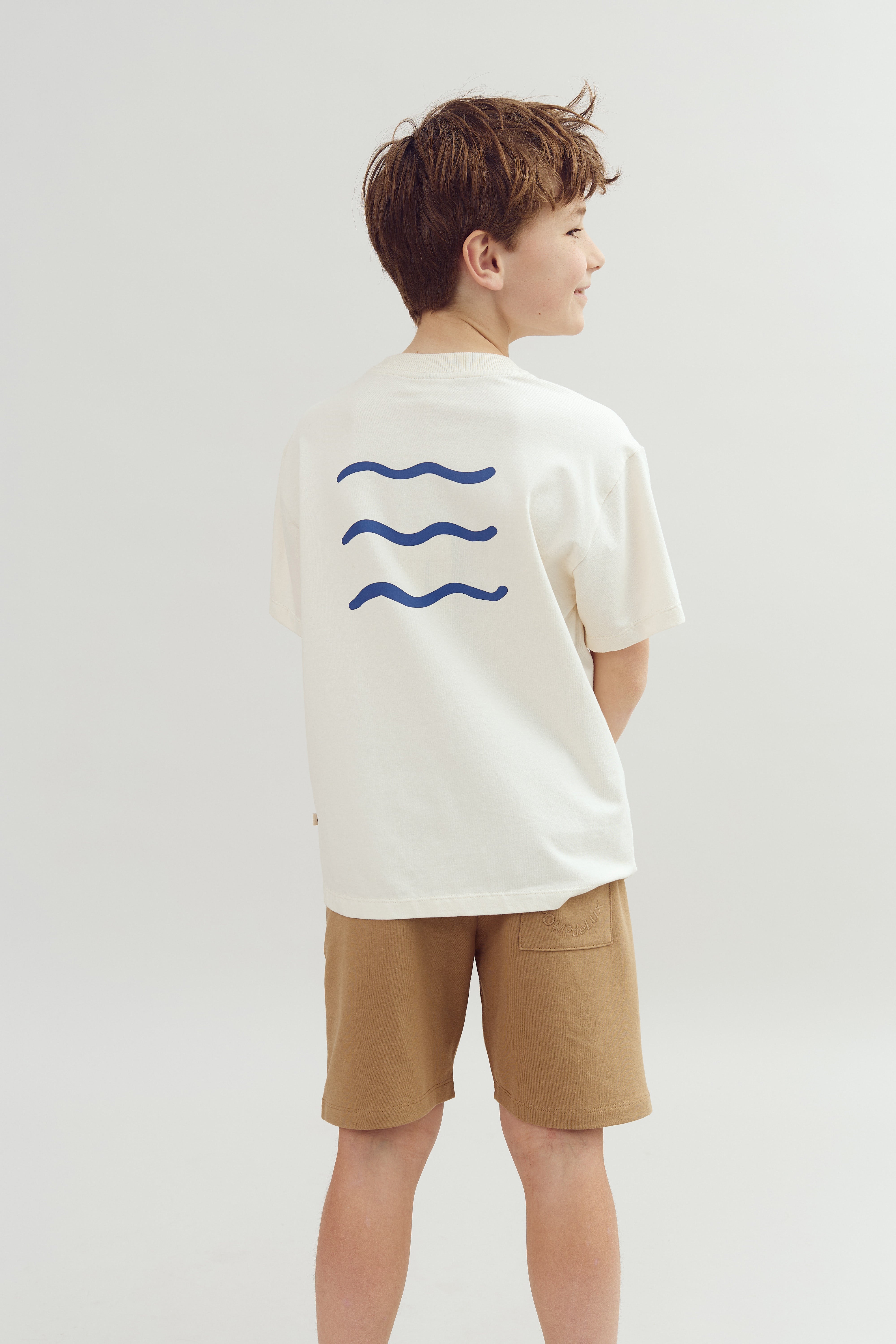 A boy wearing a white T-shirt with three blue wavy lines on the back faces sideways, standing in a neutral, plain background.