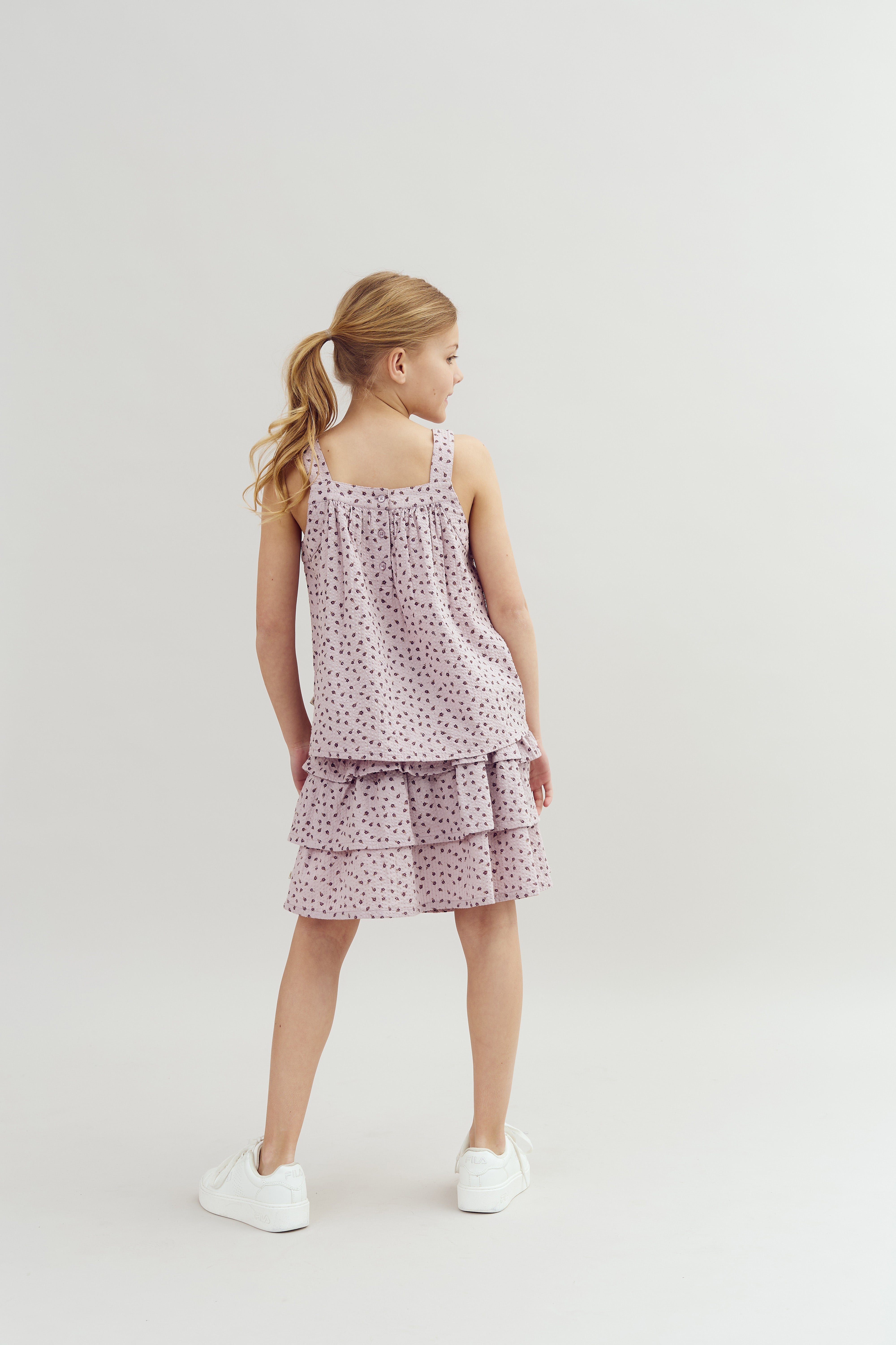A girl stands facing away, wearing a sleeveless, tiered pink dress with black polka dots and white sneakers, set against a plain white background.
