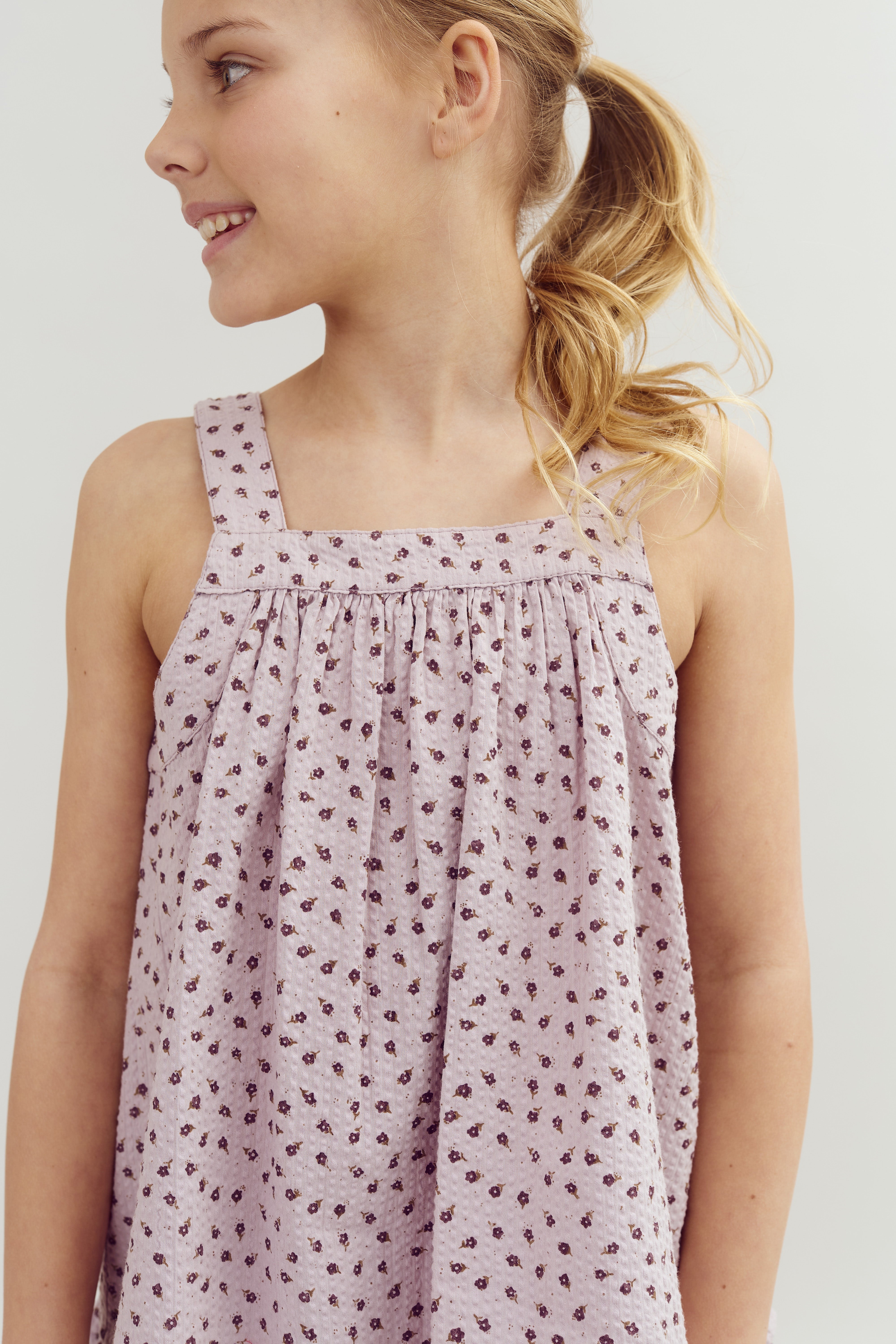 A child wearing a light purple, floral-patterned dress looks to the side, with a playful smile. Her blonde hair is styled in a ponytail, against a plain, light background.