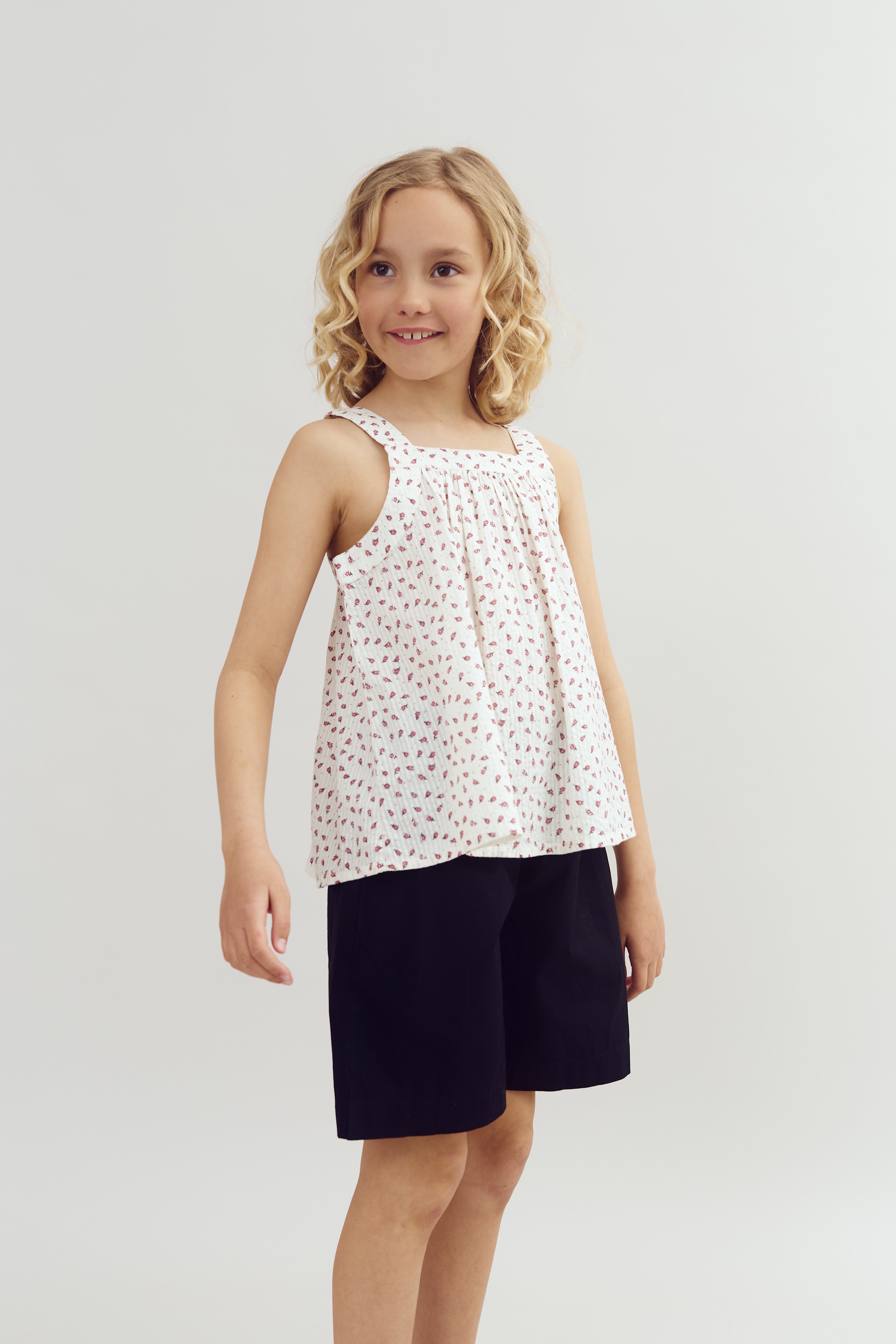 A young girl stands smiling, wearing a sleeveless, patterned white top and dark shorts, against a plain, light-colored background.