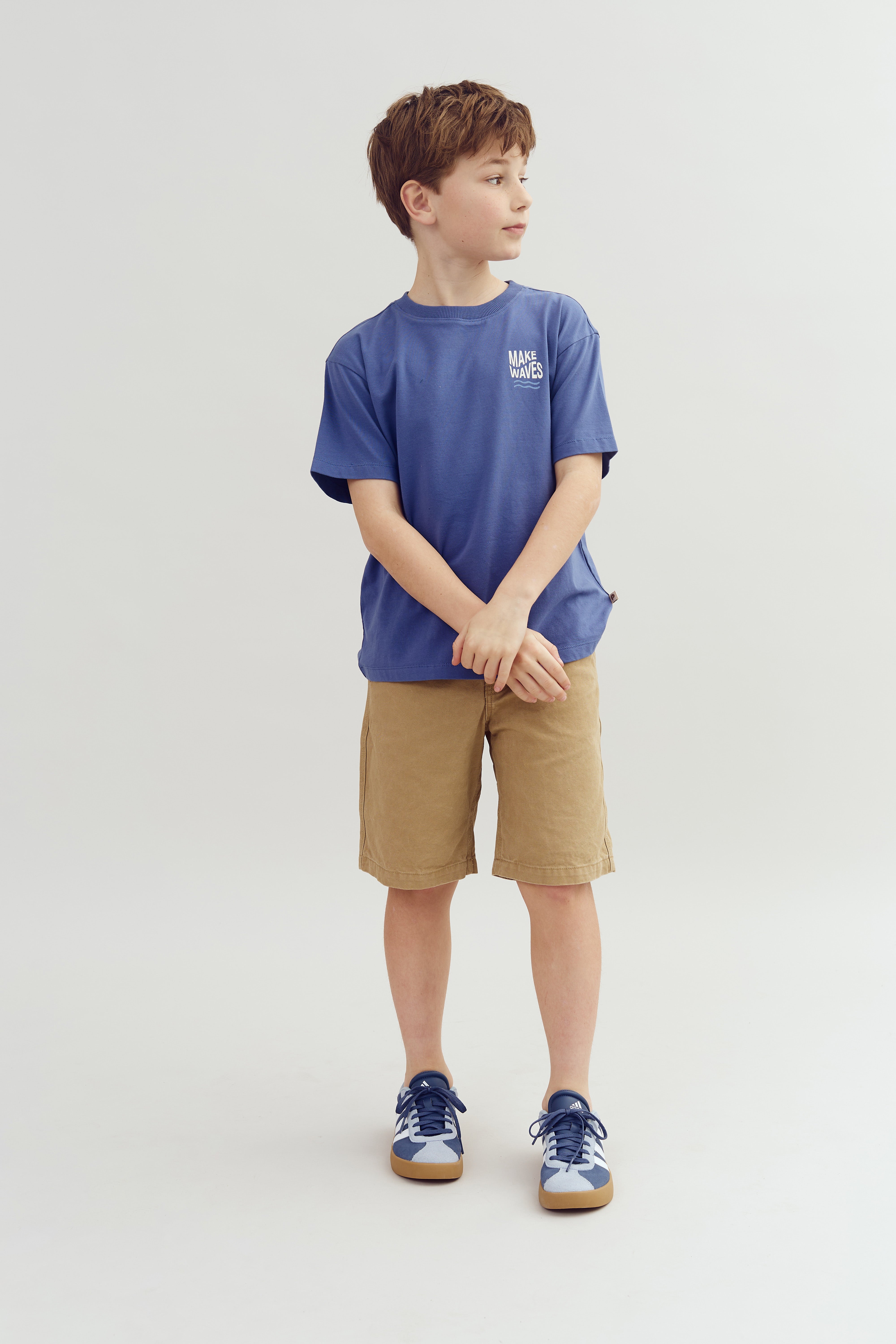 A boy in a blue shirt reads "MAKE WAVES," standing with folded hands, wearing khaki shorts and blue sneakers, against a plain light background.
