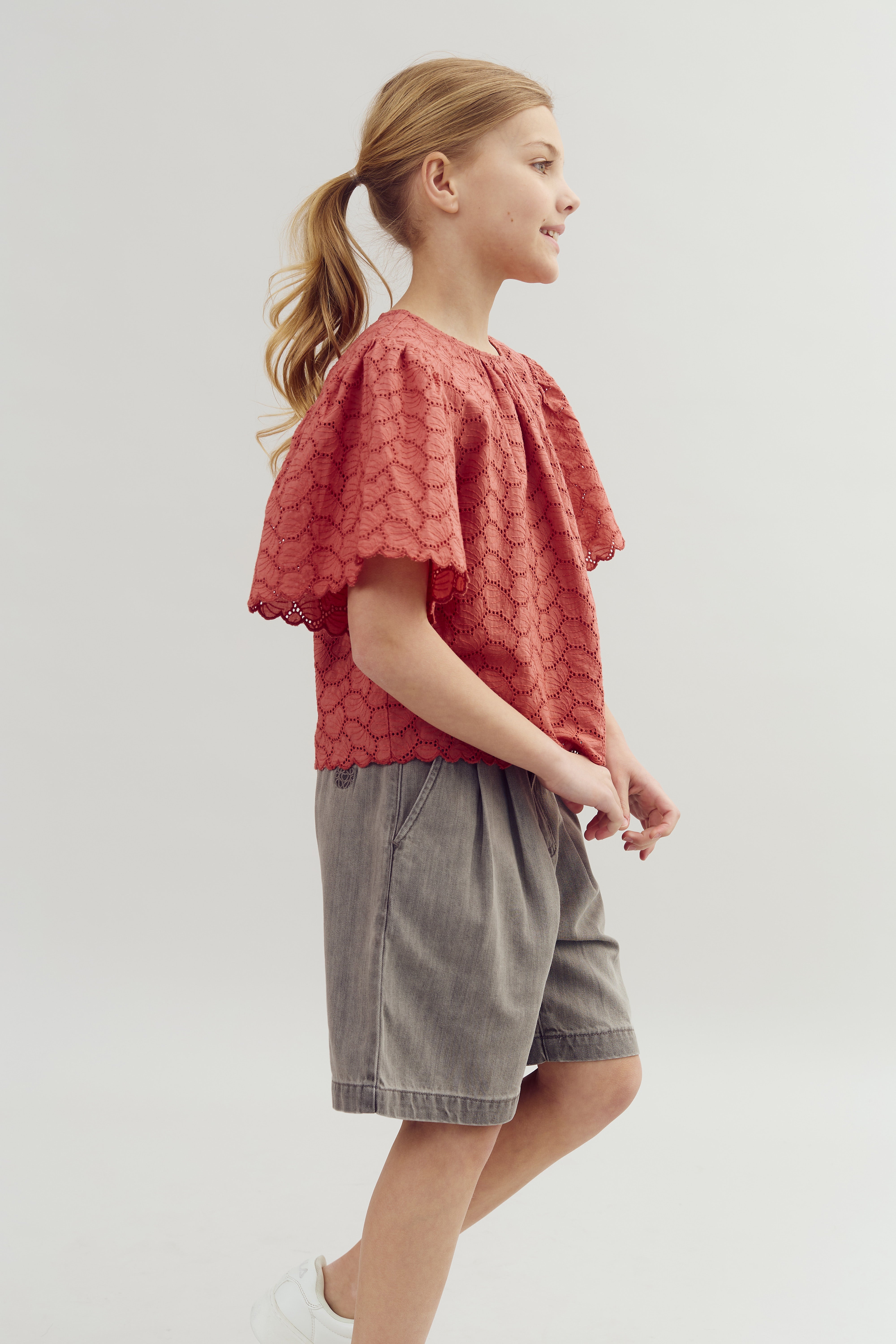 A young girl stands, smiling slightly, wearing a red scalloped top and gray shorts. Her hair is in a ponytail, and the background is a plain, light gray.