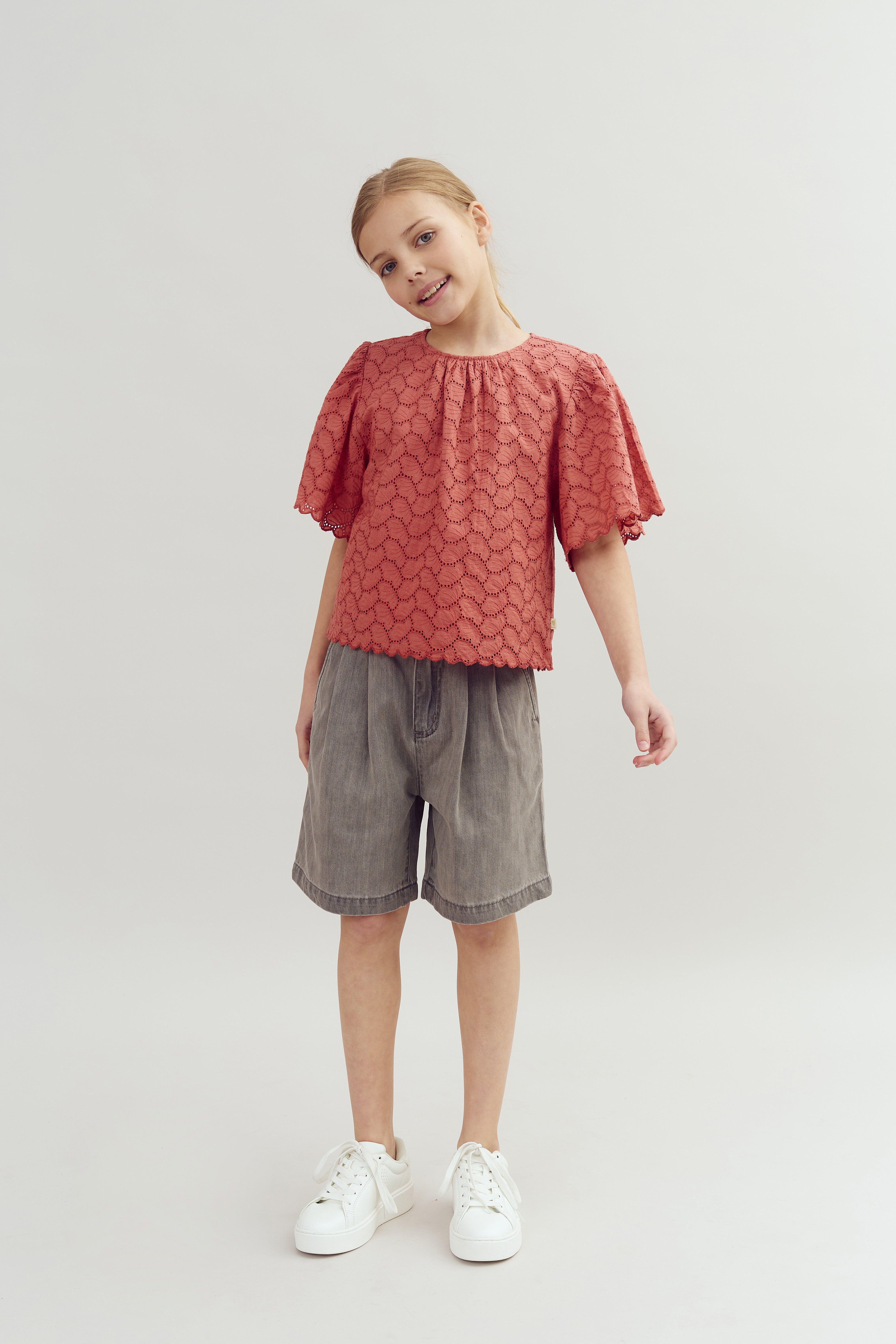 A child stands smiling, wearing a textured red top, gray shorts, and white sneakers against a plain, neutral background.
