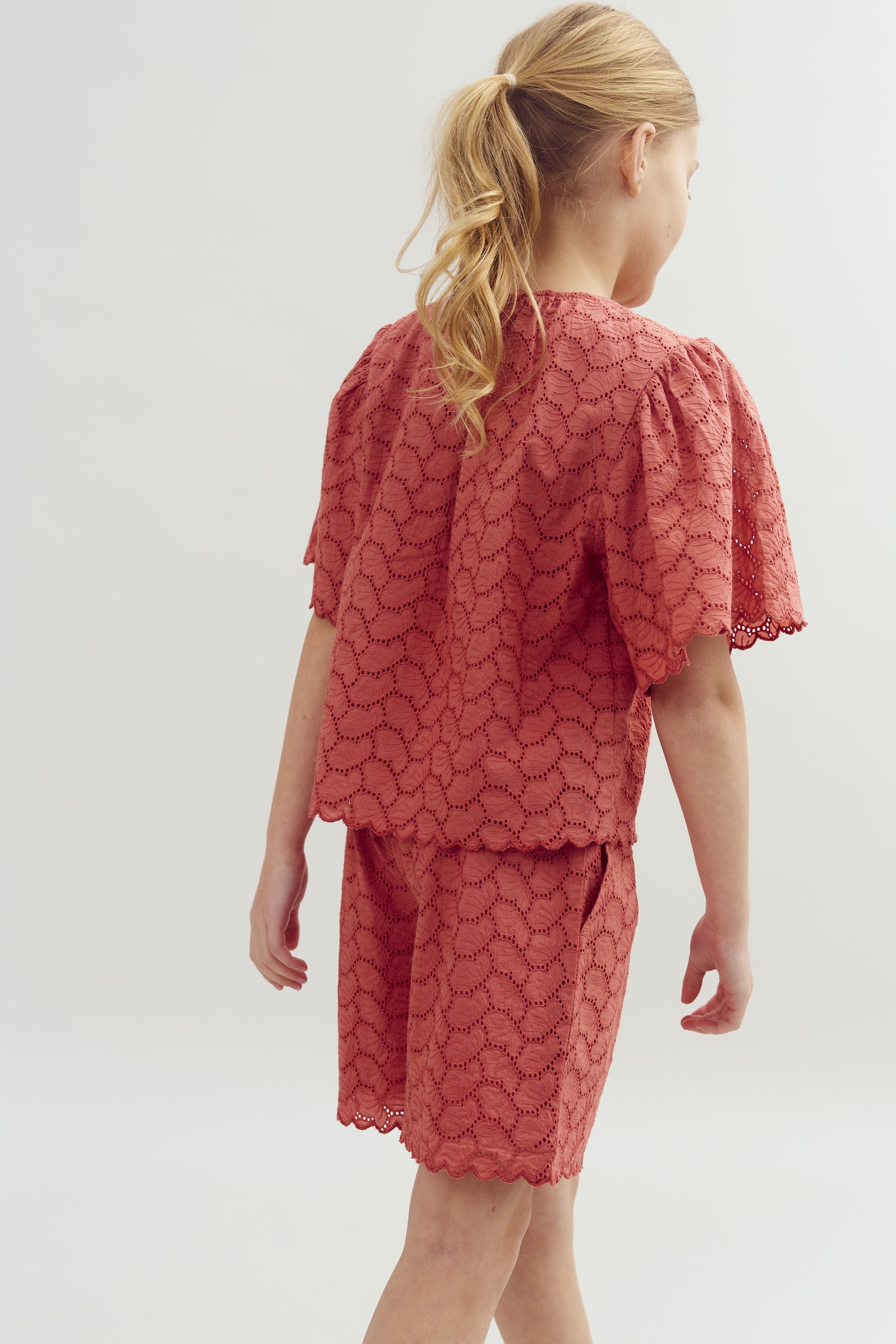 A child with blonde hair in a ponytail is wearing a pink lace outfit, consisting of a top and shorts, standing against a plain background.