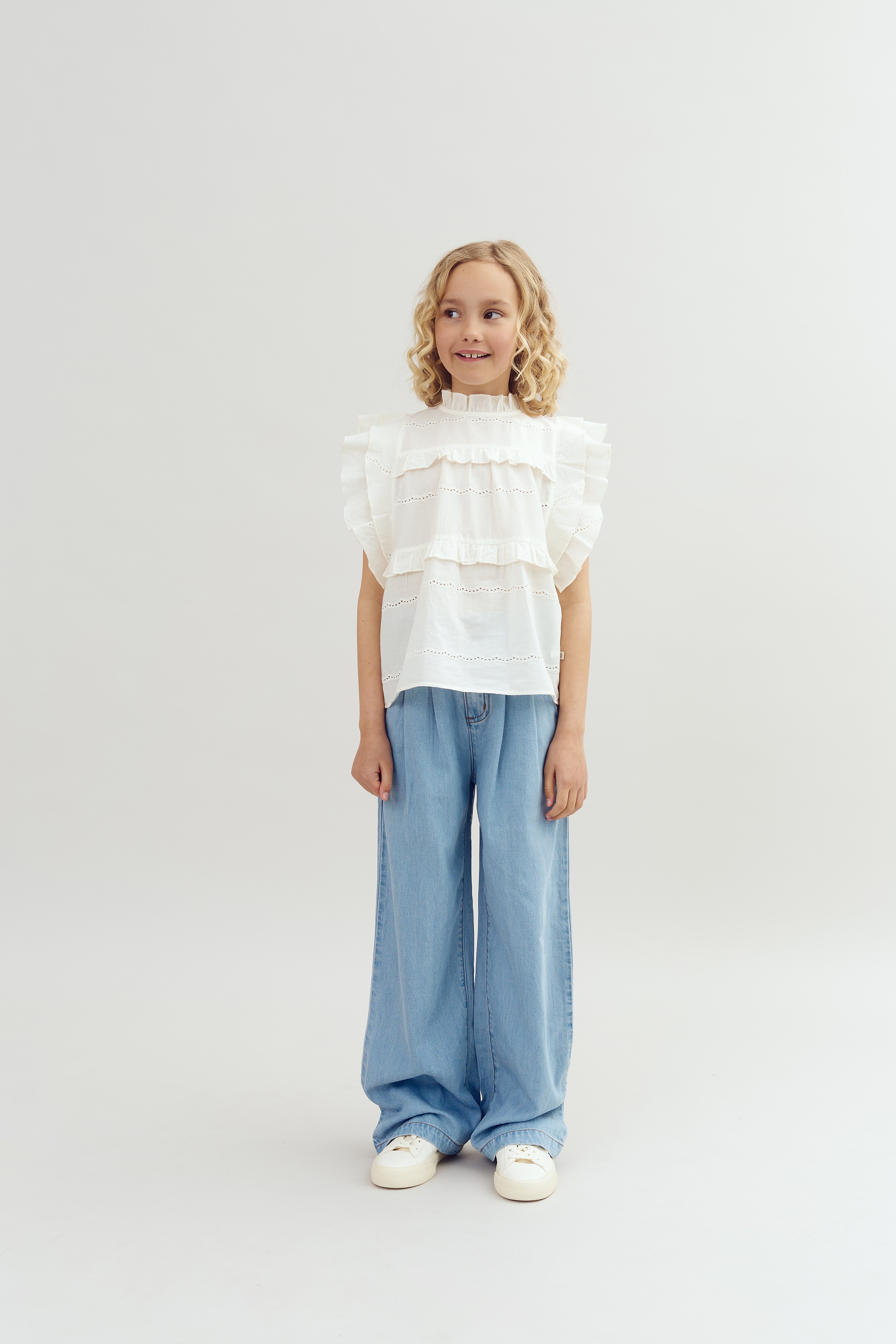 A child with curly hair stands smiling, wearing a white, ruffled blouse and wide-leg blue jeans against a plain, light-colored background.
