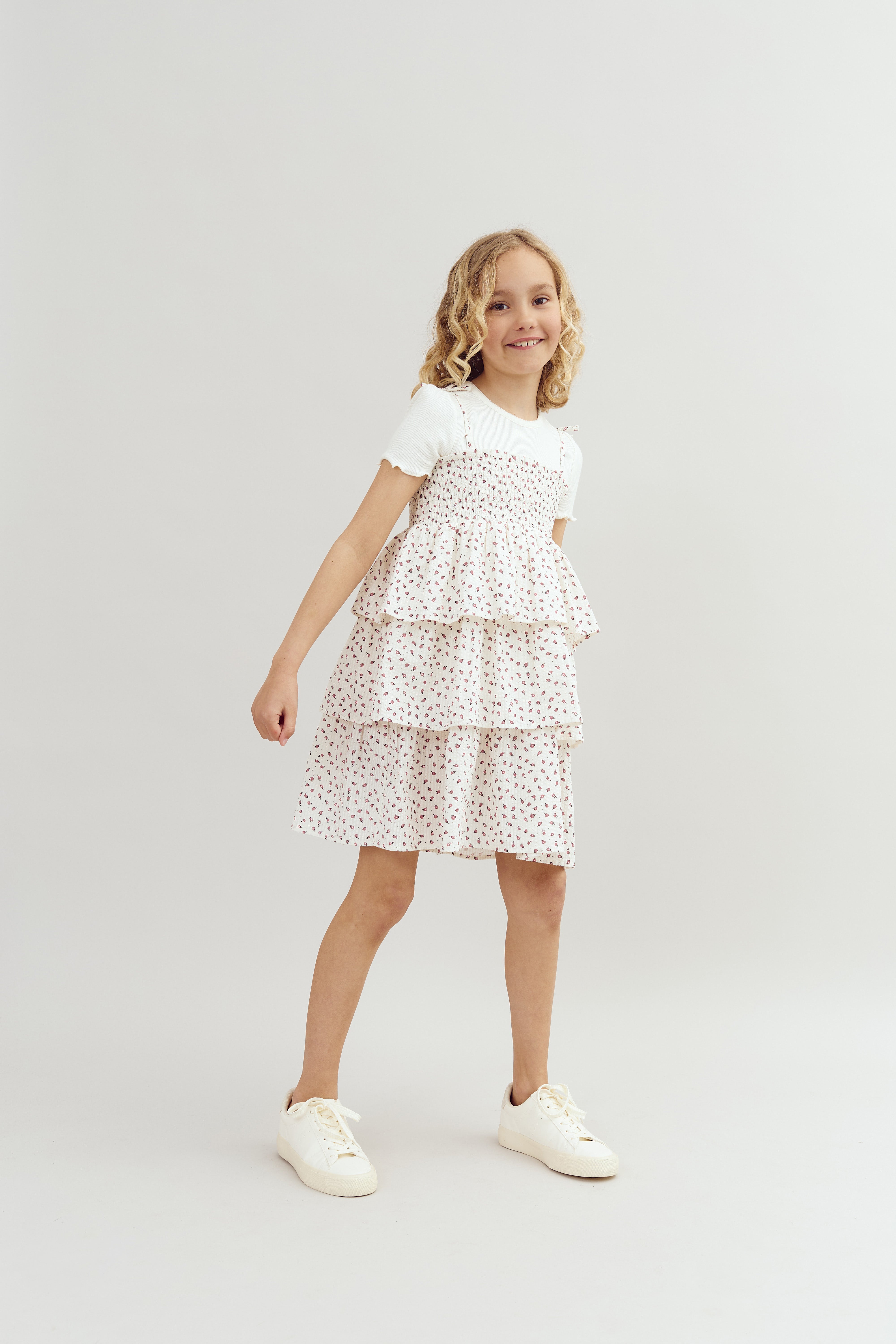 A young girl, smiling, wears a white dress with small floral patterns and ruffled layers, standing in a neutral studio setting.